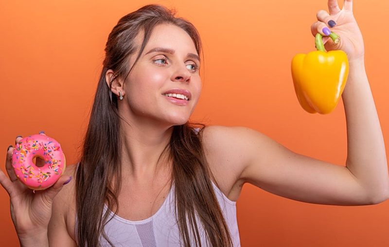 Woman in sports bra juggling choice between a donut and bell pepper on orange background.