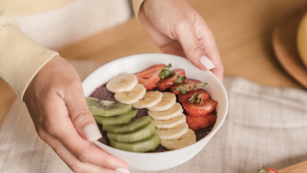 Delicious smoothie bowl topped with fresh fruits like kiwi, banana, and strawberries, held by manicured hands.