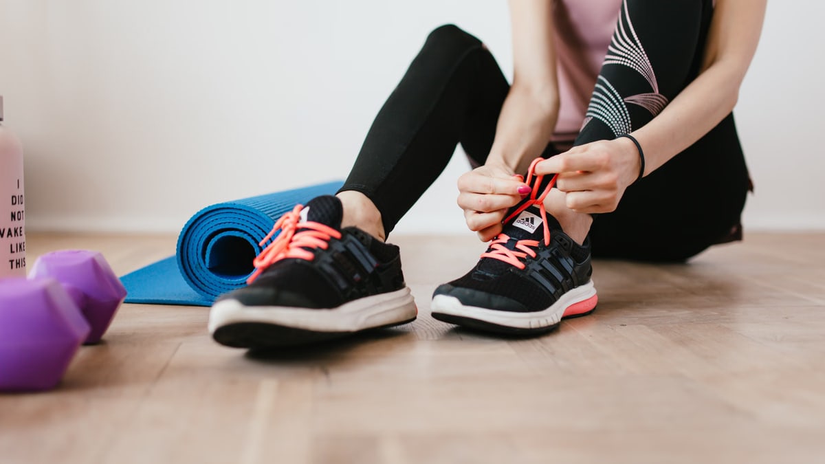 Crop sportswoman in sportswear tying sneakers while sitting on wooden floor with sport mat and dumbbells before exercising indoors